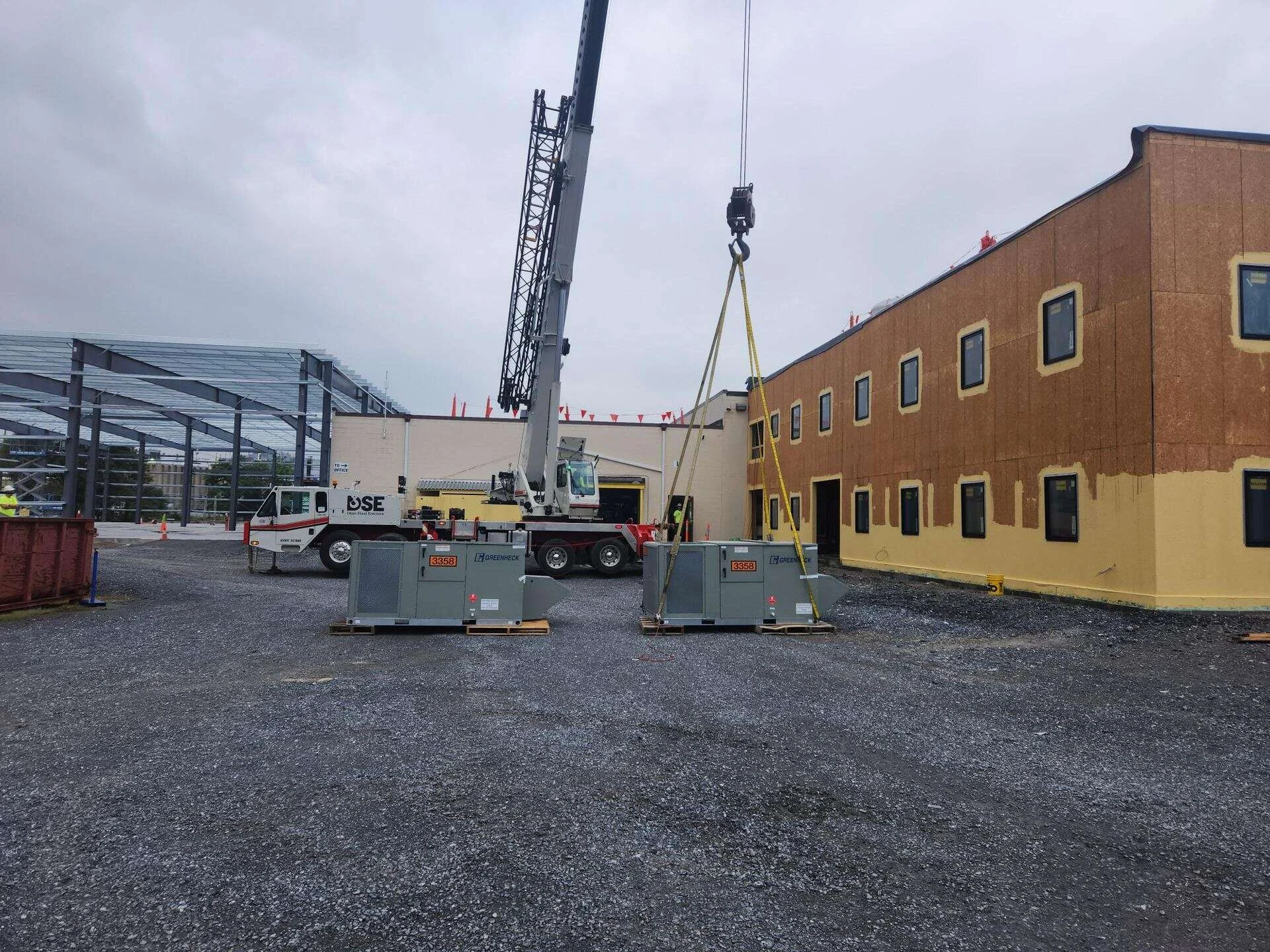 Two Greenheck packaged rooftop HVAC units staged on pallets and rigged to a crane, with the office addition and pre-engineered metal building steel frame behind them