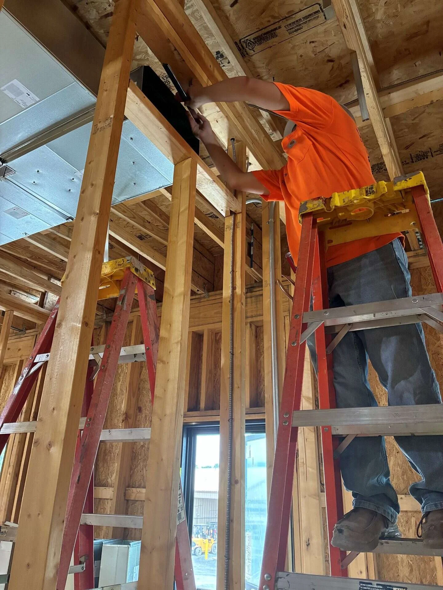 Technician on a ladder fitting sheet metal ductwork into a framed wall cavity